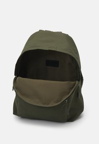 Mochila de tela verde oliva con forma redondeada, apertura con cremallera y un gran compartimento interior. Incluye una pequeña etiqueta negra en el interior.