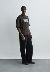 Zwarte oversized T-shirt met witte opdruk "TI AMO GCDS", gecombineerd met losse zwarte broek en zwarte schoenen; heeft een bruine pet.