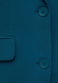 Blazer en tissu bleu sarcelle à texture lisse, doté de deux gros boutons et d'un détail de poche avant sur le côté gauche.