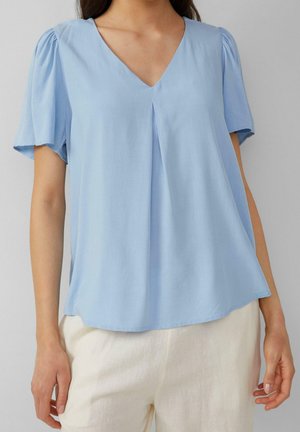 Žena v světle modré blůze s výstřihem do V a krátkými nafouklými rukávy, oblečená v krémových kalhotách, stojí před jednobarevným pozadím.