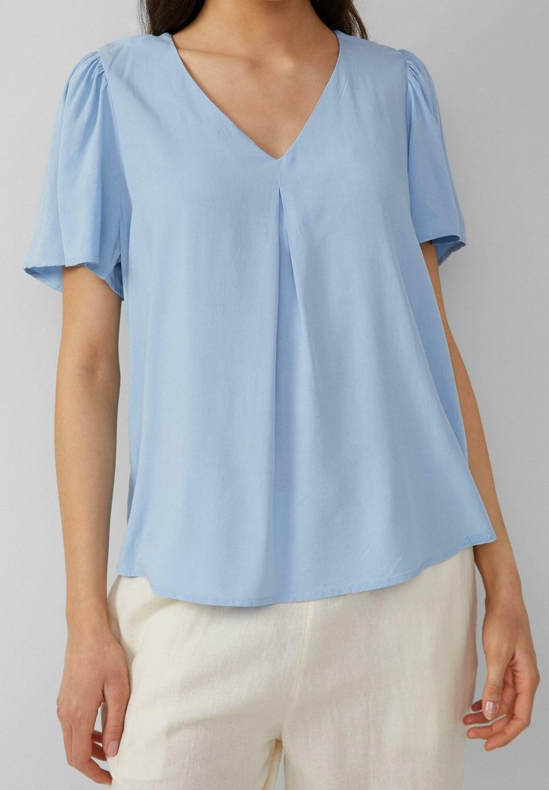 Femme portant un chemisier bleu clair à col en V avec des manches courtes bouffantes et un pantalon de couleur crème, debout devant un fond uni.