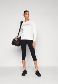 Fehér pulóver rózsaszín "BIZALOM" felirattal, fekete 3/4-es leggings, és fekete sportcipő; a modell egy fekete sporttáska tart.
