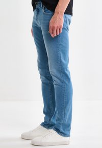 Hombre con vaqueros ajustados color azul claro y zapatillas blancas, posando frente a un fondo blanco liso.