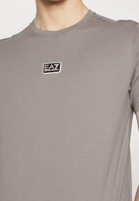 Grå bomullströja med rund halsringning. Har en svart och vit EA7 Emporio Armani logotyp på bröstet. Slät textur.