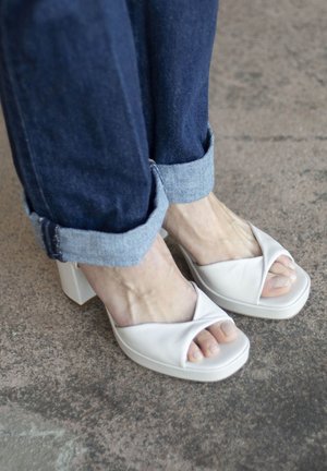 Pies con sandalias blancas de tacón abiertas en la punta, combinadas con jeans de mezclilla azul oscuro remangados, sobre una superficie texturizada.