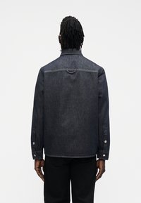 Džínová bunda v tmavě indigové barvě s klasickým límečkem, knoflíkovými manžetami a detailem zadní smyčky. Texturovaná látka se strukturálním střihem.