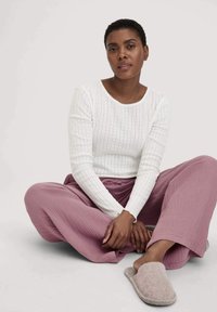 Weißes geripptes Langarmoberteil, lockere mauvefarbene Hose mit strukturiertem Muster und graue Hausschuhe. Modell sitzt mit übereinandergeschlagenen Beinen.