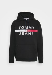 Černá mikina s kapucí vyrobená z bavlny, se zásuvným klokankovým kapsou, kapucí se stahovací šňůrkou a velkým potiskem: "TOMMY JEANS" s červenými a bílými akcenty.