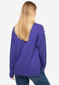 Lila gestrickter Pullover mit lockerer Passform, seitlichen Schlitzen und gerippten Bündchen. Von hinten gezeigt, kombiniert mit blauen Jeans.
