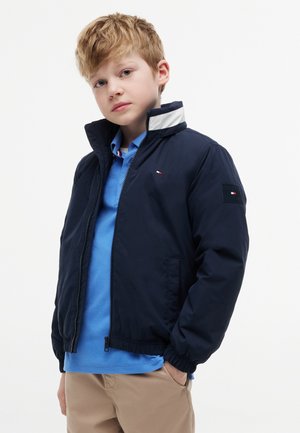 Ung dreng med lyst hår iført en navyblå jakke over en lyseblå skjorte med krave og beige bukser, stående med den ene hånd i lommen.