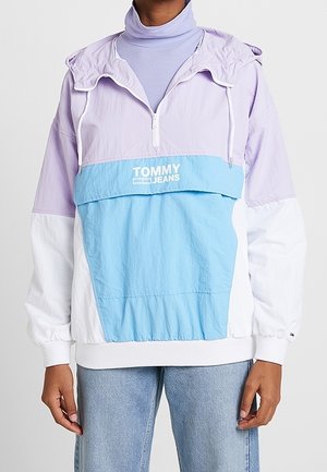 Osoba ubrana w kurtkę przeciwdeszczową Tommy Jeans w kolorach lawendy, bieli i niebieskiego, na jasnofioletowy golf i jasnoniebieskie dżinsy.