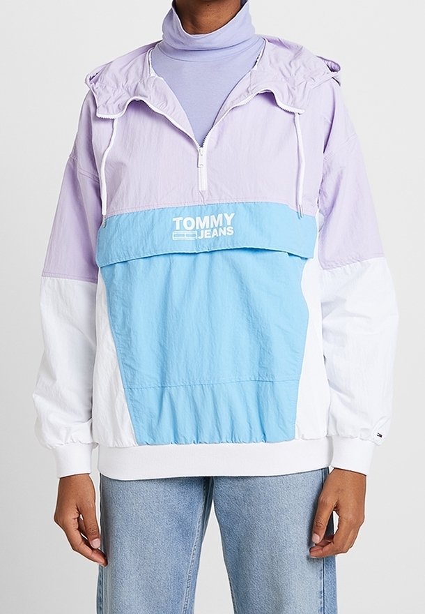 Personne portant une veste coupe-vent Tommy Jeans de couleur lavande, blanche et bleue, par-dessus un col roulé violet clair et un jean bleu clair.
