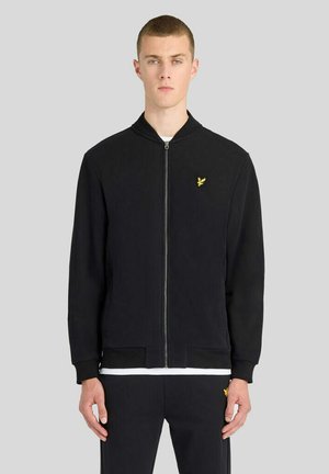 Jeune homme portant une veste noire zippée et un pantalon noir assorti avec un petit logo jaune d'oiseau sur la poitrine et la cuisse, debout devant un fond gris.