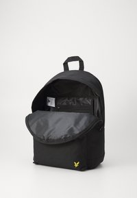 Sac à dos en tissu noir avec une grande ouverture à rabat, doté d'un intérieur lisse, d'une pochette zippée et d'un logo brodé jaune en bas.