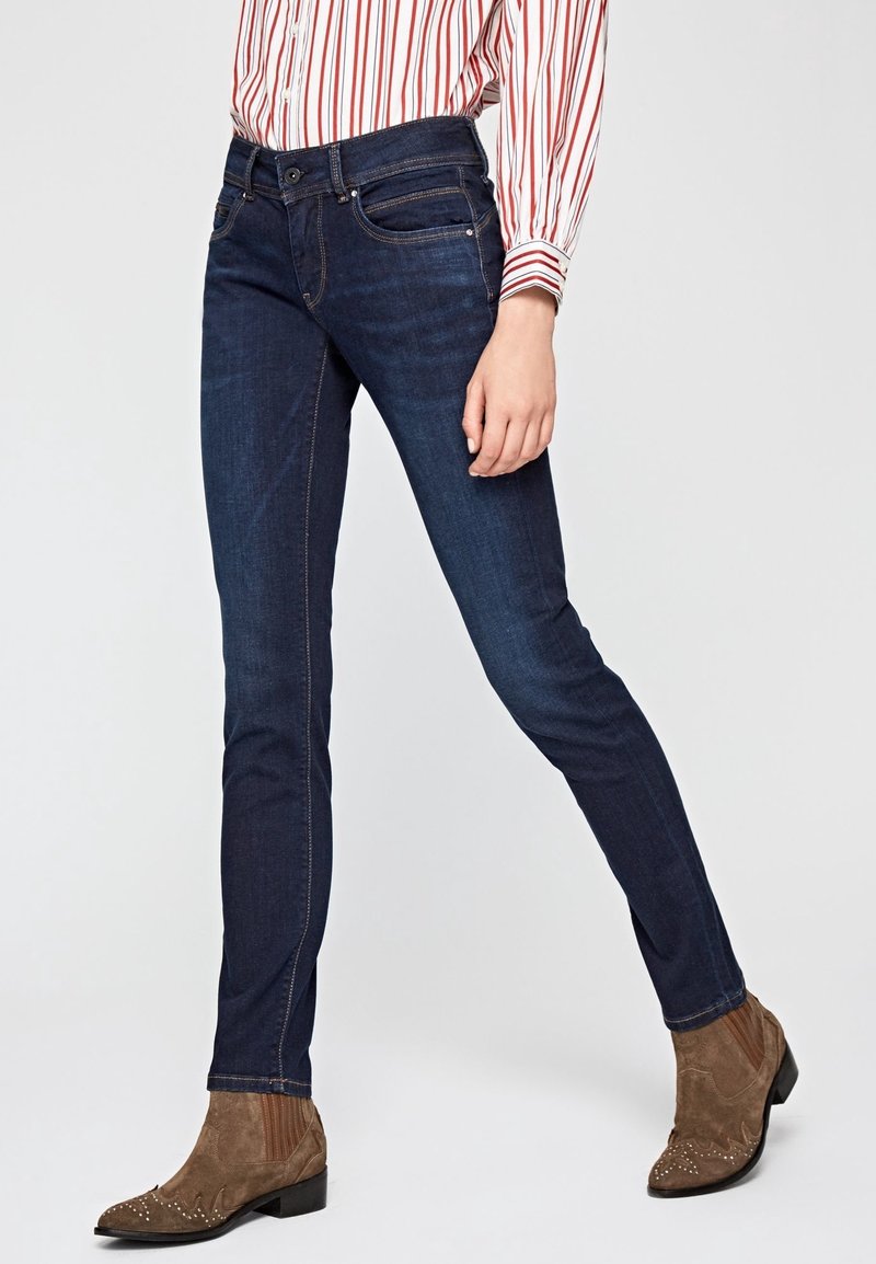 Personne portant un jean skinny bleu foncé, des bottes en daim marron avec des clous et une chemise blanche à rayures rouges verticales, debout sur un fond uni.