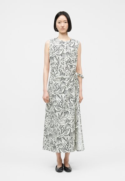 Femme aux cheveux noirs et raides portant une robe blanche sans manches avec des motifs abstraits de feuilles noires et des chaussures plates noires, debout sur fond blanc.