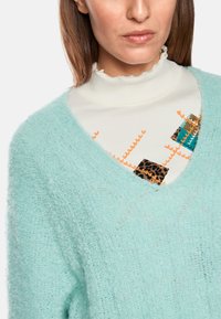 Hellblaues, flauschiges Strickpullover mit V-Ausschnitt. Darunter ein strukturiertes weißes Oberteil mit bunten Mustern, einschließlich orangefarbener und Leopardenmuster-Akzenten.