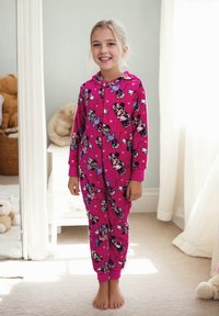 Ragazza sorridente in un morbido pigiama di pile rosa luminoso di Minnie Mouse, in piedi a piedi nudi in una camera da letto illuminata delicatamente, con adorabili orsacchiotti sullo sfondo.