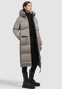 Long puffer coat in light gray with a hood, featuring a quilted design, elastic drawstring, and zippers. Worn over black attire with combat boots.