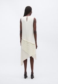 Rochie crem fără mâneci, cu tiv asimetric, având un detaliu texturat cu franjuri în partea de jos și o deschidere tip cheie la spate.