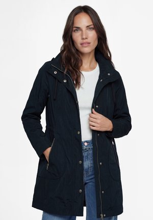 Frau mit langen braunen Haaren, die einen schwarzen Reißverschlussmantel über einem weißen Hemd und blauen Jeans trägt, steht vor einem einfarbigen weißen Hintergrund.