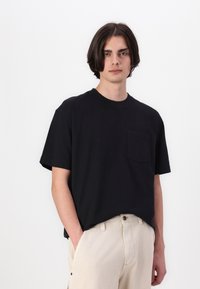 Zwarte katoenen t-shirt met ronde hals en korte mouwen, voorzien van een borstzak. Gestyled met lichtgekleurde broeken. Schoon, minimalistisch ontwerp.