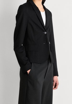 Woman wearing a fitted black blazer with two buttons over a white top, paired with loose black trousers, standing with one hand in pocket.