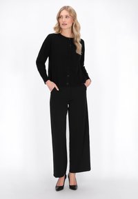 Schwarze Strickjacke mit runder Ausschnitt und Knöpfen, kombiniert mit weiten schwarzen Hosen. Das Model hat die Hände in den Taschen und trägt schwarze High Heels.