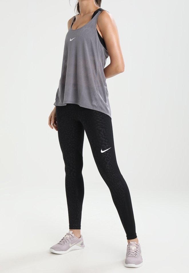 Femme debout portant un haut sans manches gris Nike, des leggings Nike noirs à motifs, et des baskets Nike violet clair sur un fond blanc uni.