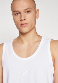 Biely tielko s okrúhlym výstrihom a malým vyšitým logom Nike na hrudi. Hladká textúra látky, minimalistický dizajn.