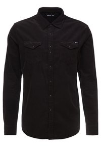 Camisa de pana negra con cuello abotonado, dos bolsillos en el pecho y una sutil marca en el bolsillo, con un acabado de tejido texturizado.