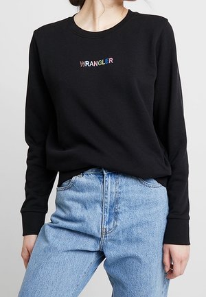 Nainen pukeutunut mustaan pitkähihainen paitaan, jossa monivärinen teksti "WRANGLER", ja vaaleansinisiin korkeavyötäröisiin farkkuihin, seisoo yhdellä kädellä sivulla.