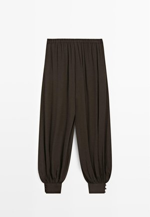 Losse donkerbruine broek met elastische tailleband en geplooide boorden met knoopdetails bij de enkels, afgebeeld op een witte achtergrond.