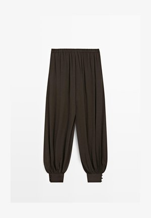 Løse mørkebrune bukser med elastisk talje og opsamlede manchetter med knapdetaljer ved anklerne, vist på hvid baggrund.