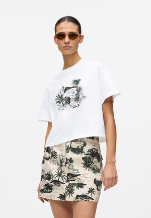 Femme portant des lunettes de soleil rondes, un t-shirt blanc imprimé de palmiers et de rhinocéros, et une jupe beige à motif tropical.