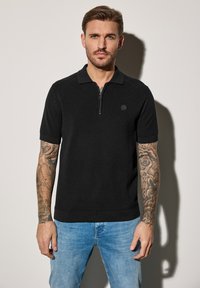 Svart kortärmad polo-tröja med dragkedja och texturerad tyg. Har en rund logotypapplikation på bröstet och en rak fåll.