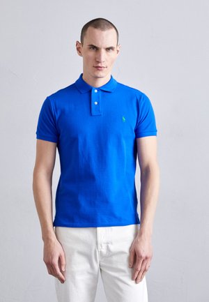 Polo shirt bleu en coton, avec un col classique, deux boutons et un petit logo vert sur la poitrine gauche. Porté avec un pantalon blanc.