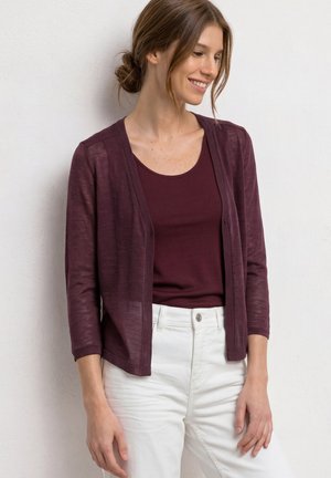 Femme aux cheveux bruns attachés en chignon, portant un cardigan violet foncé sur un top assorti et un pantalon blanc, debout contre un mur clair.