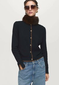 Cardigan lavorato a maglia blu navy con rifiniture marroni, featuring un colletto in pelliccia sintetica e bottoni dorati. Indossato con jeans a vita alta blu chiaro.