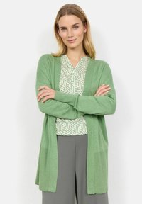 Ljusgrön cardigan med ribbat mönster, öppen fram, och sidofickor, lager på lager över en mönstrad topp och grå byxor.