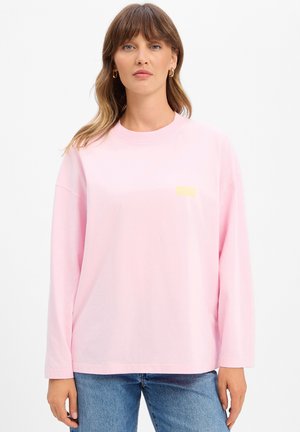 Frau mit schulterlangem braunem Haar, die goldene Creolen, ein lockeres hellrosa Langarmshirt und blaue Jeans trägt, steht vor weißem Hintergrund.