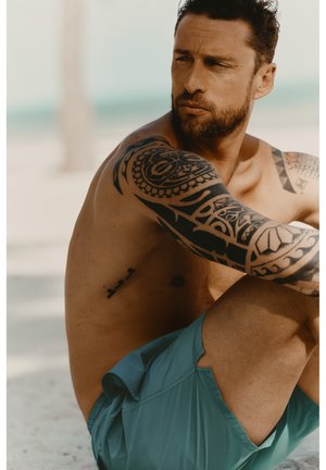 Skjorteløs mand med stamme-tatoveringer på armen, der sidder udendørs iført grønne shorts og ser eftertænksomt til siden.