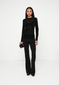 Top preto ajustado de mangas longas com uma textura transparente, combinado com calças pretas de cintura alta e sapatos de salto fino pretos, segurando uma pequena mala preta.