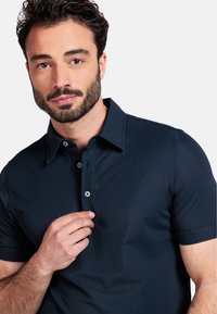 Polo shirt marine avec un col classique, des manches courtes et trois boutons. Texture lisse avec un léger éclat, comportant une poche unique sur la poitrine.