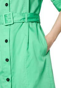 Grünes Hemdkleid mit tailliertem Gürtel, kurzen Ärmeln, durchgehender Knopfleiste und Seitentaschen. Glatte Textur mit einer einfarbigen Oberfläche.