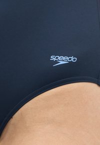 Κοντινή λήψη υφάσματος μαγιό Speedo σε μαύρο χρώμα με εκτυπωμένο λογότυπο πάνω σε μαυρισμένο δέρμα κοντά στη μέση ενός ατόμου.