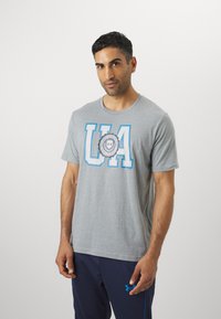 Grå bomulds t-shirt med rund halsudskæring. Har iøjnefaldende blå "UA" bogstaver og et rundt logo i midten. Korte ærmer, afslappet pasform.