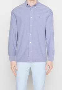 Muž v světle modré texturované košili na knoflíky s dlouhými rukávy a světle modrých kalhotách, stojící proti jednoduchému pozadí.