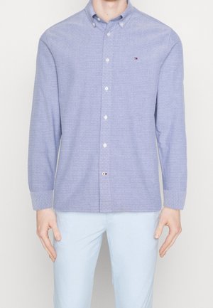 Mann in einem hellblauen, strukturierten Hemd mit langen Ärmeln und hellblauen Hosen, der vor einem schlichten Hintergrund steht.