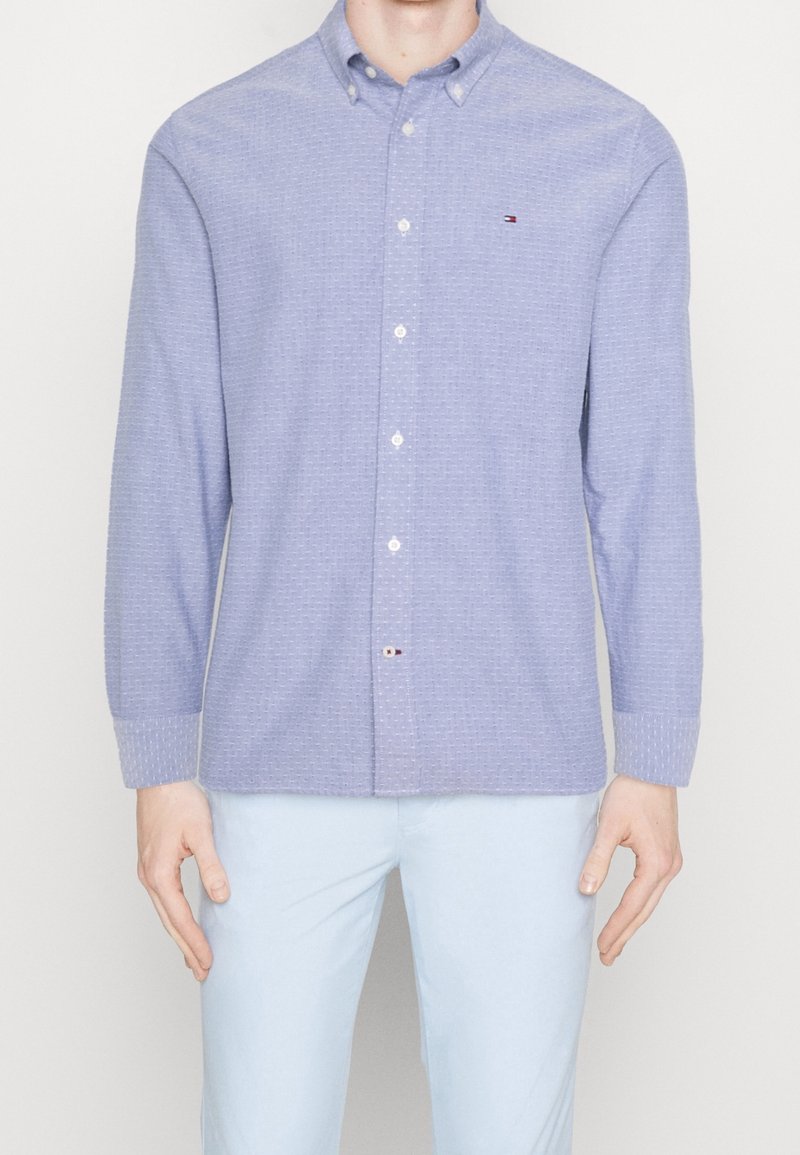 Mies, joka päälleen pukeutuneena vaaleansiniseen textuuri-paitaan, jossa on pitkät hihat, ja vaaleansinisiin housuihin, seisoo tasaisen taustan edessä.